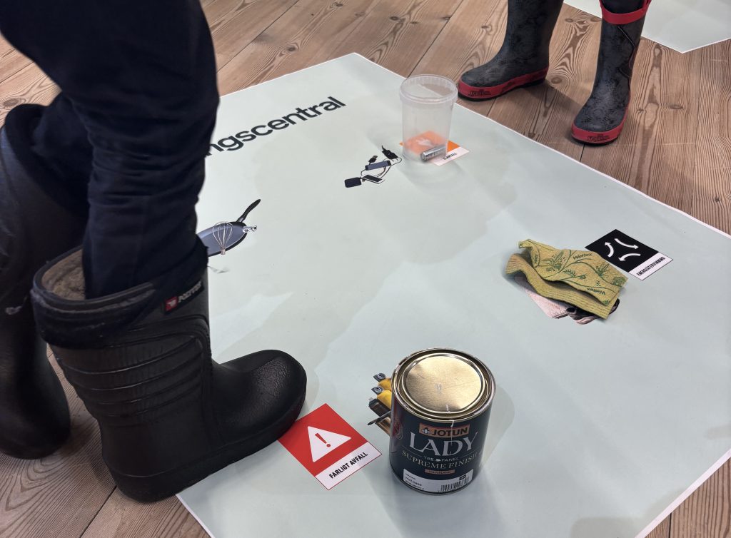 Foto. Sortering av fyra sorters avfall på en affisch på golvet med närbild på två barns fötter.